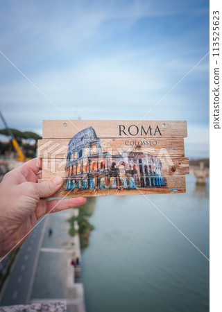 Hand holding Rome Colosseum postcard, designed as wood piece with ROMA and COLOSSEO . Slight blurry cityscape background, river, bridge, partly cloudy sky possibly Rome. 113525623