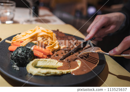 Close up view of gourmet meal on black plate with fettuccine pasta, cooked veggies, and steak in peppercorn sauce. Restaurant setting with warm lighting. Close up view of gourmet meal on black plate with fettuccine pasta, cooked veggies, and steak in peppercorn sauce. Restaurant setting with warm lighting. 113525637