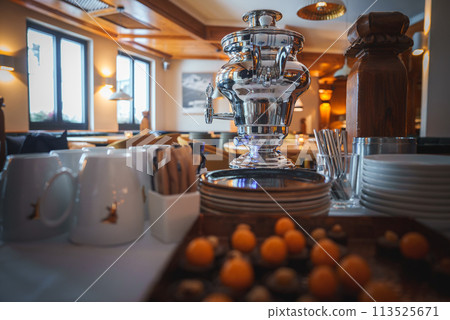Elegant buffet setup with shiny silver samovar, white plates, wooden pepper mill, ceramic mugs, wooden utensils, kumquats, warm interior atmosphere. Ideal for meal service. Elegant buffet setup with shiny silver samovar, white plates, wooden pepper mill, ceramic mugs, wooden utensils, kumquats, warm interior atmosphere. Ideal for meal service. 113525671