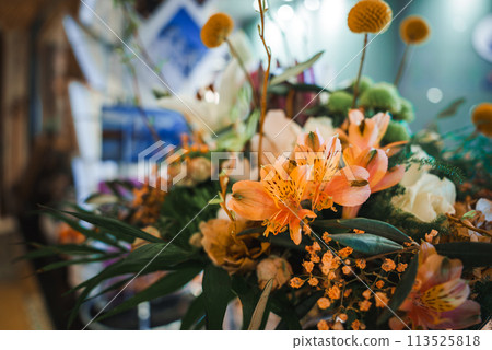 Colorful floral arrangement featuring vibrant orange lilies, small orange flowers, yellow button like blooms, and varied greenery in a blurry indoor setting. 113525818