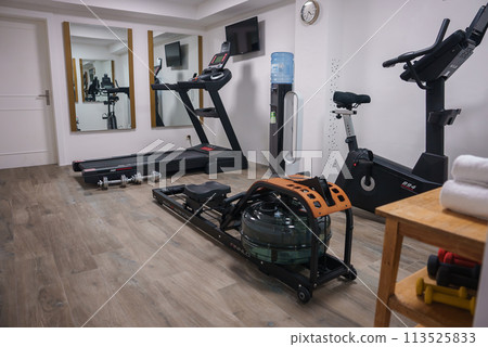 Modern home gym with various exercise machines rowing machine, treadmill, dumbbells, exercise bike. Wall clock, water cooler, mirrors. Private fitness space. Modern home gym with various exercise machines rowing machine, treadmill, dumbbells, exercise bike. Wall clock, water cooler, mirrors. Private fitness space. 113525833