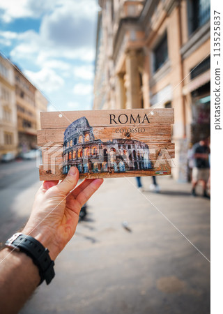 Hand holding souvenir with Colosseum depiction, ROMA and COLOSSEO printed on wood. Background of Rome street with people, European buildings. Sky partly cloudy. Main focus is on souvenir. Hand holding souvenir with Colosseum depiction, ROMA and COLOSSEO printed on wood. Background of Rome street with people, European buildings. Sky partly cloudy. Main focus is on souvenir. 113525837