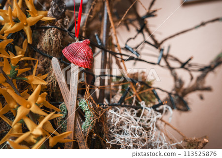 Close up view of textured red knitted hat hanging from ribbon, surrounded by dried plants, twigs, and yellow star shaped flowers in rustic, natural themed arrangement. Indoor display. Close up view of textured red knitted hat hanging from ribbon, surrounded by dried plants, twigs, and yellow star shaped flowers in rustic, natural themed arrangement. Indoor display. 113525876