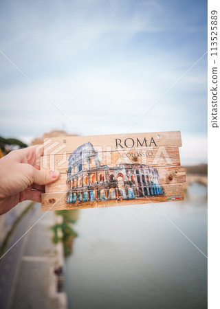 Hand holding wooden postcard with Colosseum illustration from Rome, Italy. ROMA and COLOSSEO displayed, rustic design on wood background near river and bridge. 113525889