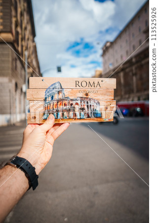 Hand holding souvenir sign with Colosseum image in Rome. ROMA and Colosseo displayed. Blurred street scene background. Urban setting, partly cloudy. Hand holding souvenir sign with Colosseum image in Rome. ROMA and Colosseo displayed. Blurred street scene background. Urban setting, partly cloudy. 113525926