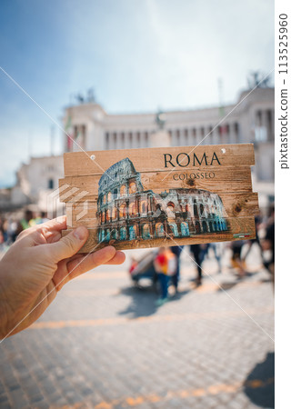 Hand holding Colosseum illustration postcard with ROMA and COLOSSEO in Rome, Italy. Blurred urban background aligns with street architecture, sunny day vibes. Hand holding Colosseum illustration postcard with ROMA and COLOSSEO in Rome, Italy. Blurred urban background aligns with street architecture, sunny day vibes. 113525960