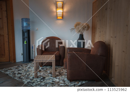Cozy corner in room with warm, rustic ambiance. Light walls contrast wood paneling. Leather armchairs, wicker table, water cooler. Decorative vase, elegant lighting. Comfortable and tranquil space. 113525962
