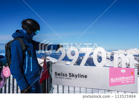 Person in winter ski attire stands by 2976 Swiss Skyline Eiger, Monch, Jungfrau sign at Murren ski resort, Switzerland, with panoramic Swiss Alps view. 113525984