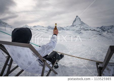 Winter scene at Zermatt ski resort person in snow gear holds up beer in deck chair overlooking Matterhorn mountain. Perfect blend of adventure and relaxation. Winter scene at Zermatt ski resort person in snow gear holds up beer in deck chair overlooking Matterhorn mountain. Perfect blend of adventure and relaxation. 113525985