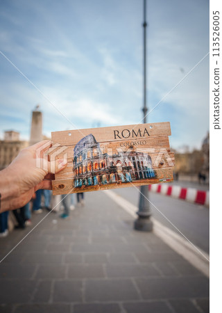 Hand holding wooden postcard with Colosseum illustration in Rome, Italy. Playful alignment with urban background. No clear landmarks but suggests Roman setting. 113526005