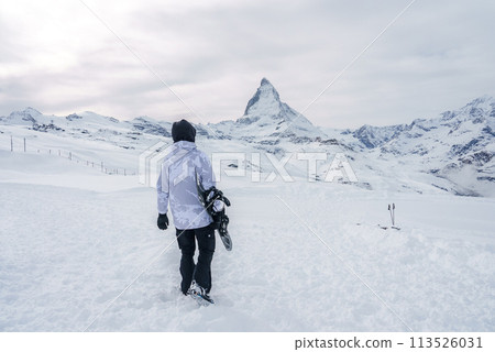 Snowboarder in camouflage winter gear walking towards Matterhorn peak in Zermatt ski resort, Switzerland. Serene snow covered landscape with iconic mountain silhouette. 113526031
