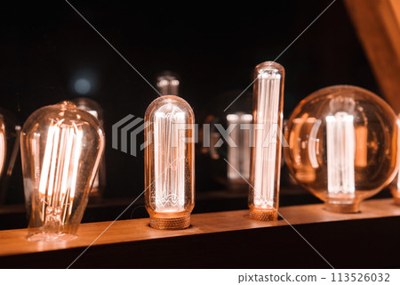 Collection of various shaped illuminated light bulbs emitting warm, amber glow on wooden surface. Filaments resemble squirrel cage type. Dark background creates cozy atmosphere. Location Unknown. 113526032