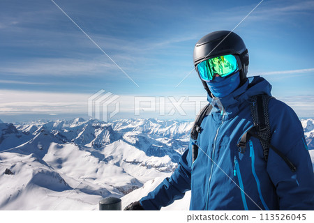 Person in winter sports attire stands against snow covered Swiss Alps backdrop, wearing blue ski jacket, helmet, and goggles. Scene likely in Murren ski resort, showcasing alpine landscape. 113526045