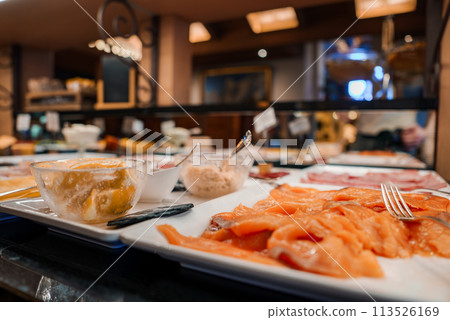 Buffet spread with citrus dessert and sliced salmon on white plate in warm, inviting restaurant or dining area with out of focus background dishes and bar area. Buffet spread with citrus dessert and sliced salmon on white plate in warm, inviting restaurant or dining area with out of focus background dishes and bar area. 113526169