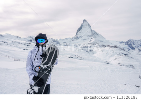 Snowboarder in winter gear holding black snowboard in snowy landscape near Zermatt, Switzerland. Matterhorn mountain in background under overcast sky. 113526185