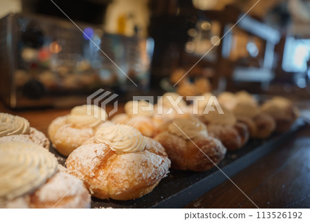 Close up view of cream filled pastries dusted with powdered sugar, decorated with a swirl of cream, neatly arranged on a dark tray. Background reveals a cozy bakery or cafe interior. 113526192