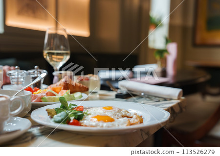 Luxurious breakfast setting in high end hotel in Rome. Close up of eggs, tomatoes, leafy greens on white plate. Glass of white wine in background. Classic elegant design. Luxurious breakfast setting in high end hotel in Rome. Close up of eggs, tomatoes, leafy greens on white plate. Glass of white wine in background. Classic elegant design. 113526279