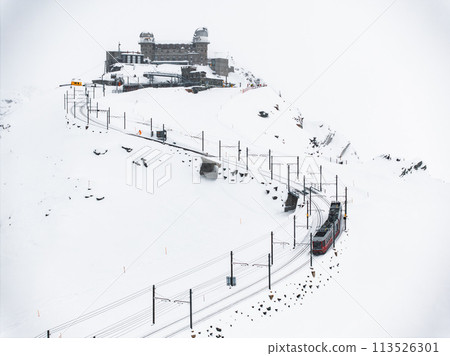 Aerial shot shows a red and white Swiss train climbing Zermatt's snowy slopes, Switzerland. Tracks cut through snow, revealing rocks and a dome building against a cloudy sky. 113526301