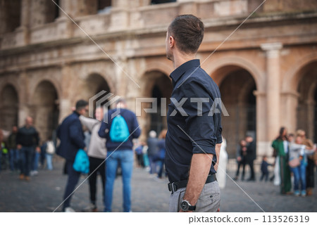 Man in dark shirt and light pants faces ancient building with arches in bustling public square, possibly Europe. Casual, touristic scene in unknown location. 113526319