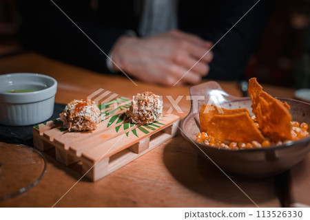 Close up of a wooden serving board with coconut covered treats, green liquid bowl, and ceramic plate of orange crisps and nuts in a cozy dining space. 113526330