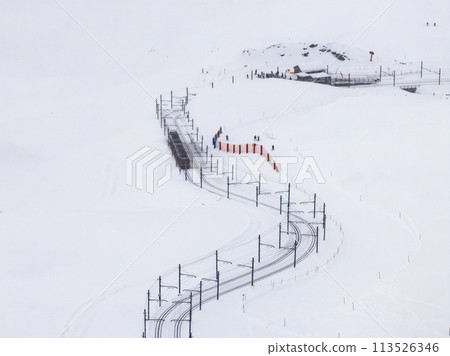 Aerial view of a snowy landscape in Zermatt, Switzerland, featuring a train track winding up the mountain and a group of people near a station, highlighting the serene beauty of the winter resort. 113526346