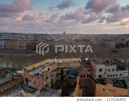 Rome from above at dawn or dusk, with terracotta roofs and the winding Tiber River. St. Peter's Basilica's dome stands out against a pink and blue sky. Rome from above at dawn or dusk, with terracotta roofs and the winding Tiber River. St. Peter's Basilica's dome stands out against a pink and blue sky. 113526403