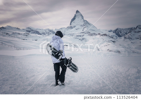 Snowboarder at Zermatt ski resort, facing Matterhorn mountain. Dressed in winter gear and holding a snowboard with bindings. Serene and contemplative scene with anticipation for adventure. Snowboarder at Zermatt ski resort, facing Matterhorn mountain. Dressed in winter gear and holding a snowboard with bindings. Serene and contemplative scene with anticipation for adventure. 113526404