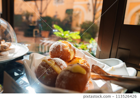 Plate of freshly baked sugared pastries, like Italian bomboloni with cream or jam filling. Set on a table in the courtyard of a luxury hotel in Rome, with golden brown exterior and creamy centers. 113526426
