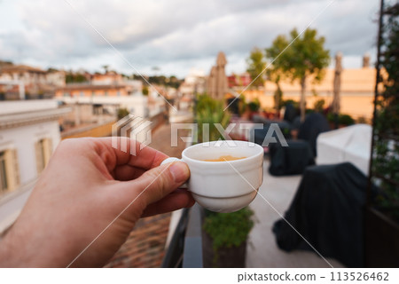 Luxury terrace in Rome close up of hand holding white espresso cup, rooftops of warm colored buildings in the background, hints of greenery and overcast sky. Relaxing atmosphere. Luxury terrace in Rome close up of hand holding white espresso cup, rooftops of warm colored buildings in the background, hints of greenery and overcast sky. Relaxing atmosphere. 113526462