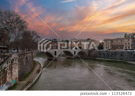 Urban river scene with stone bridge spanning grey water under an overcast sky. Trees line one bank, European buildings on the other, parked cars on street. Urban river scene with stone bridge spanning grey water under an overcast sky. Trees line one bank, European buildings on the other, parked cars on street. 113526472