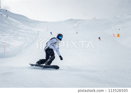 Snowy mountain landscape with winter sports activity. Snowboarder in white jacket and black pants carving turn on slope with snowy spray. Ski resort scene with skiers and lift. Snowy mountain landscape with winter sports activity. Snowboarder in white jacket and black pants carving turn on slope with snowy spray. Ski resort scene with skiers and lift. 113526505