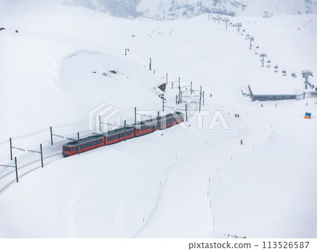 Aerial shot of busy Zermatt ski resort, with a red train on snowy tracks and a ski lift going up the mountain. Small skiers dot the scene, enjoying winter sports on a chilly, cloudy day. 113526587