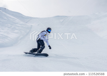 Snowboarder in white jacket navigating snowy slope with black pants, gloves, leaning into a turn. Background shows mountainous terrain under overcast sky. 113526588