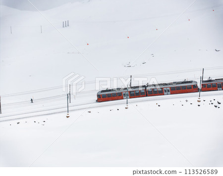 Aerial shot a bright red train glides through snowy Zermatt, beside a lone figure walking near the tracks. Sparse ski signs and red flags dot the serene landscape. Aerial shot a bright red train glides through snowy Zermatt, beside a lone figure walking near the tracks. Sparse ski signs and red flags dot the serene landscape. 113526589