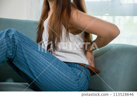 Asian beautiful young woman touching back pain sitting in sofa she suffering from backache at home, female holding her lower back feeling painful waist hurt massage in living room, healthcare medical 113528646