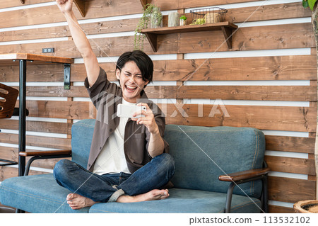 A young Asian man watching sports on his smartphone while relaxing at home (fist pump, happy) A young Asian man watching sports on his smartphone while relaxing at home (fist pump, happy) 113532102