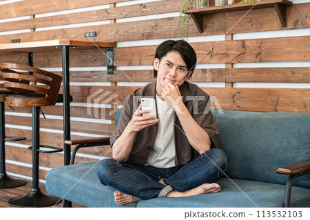 A young man relaxing at home using a smartphone (considering, thinking, having trouble, worrying) A young man relaxing at home using a smartphone (considering, thinking, having trouble, worrying) 113532103