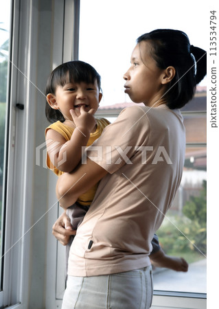 Asian Woman Holding her Baby in her Arms While Standing Next to a Window at Home. Asian Woman Holding her Baby in her Arms While Standing Next to a Window at Home. 113534794