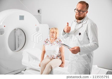 Medical computed tomography or MRI scanner. Doctor holding results of MRI, showing thumbs up gesture. Woman patient sitting on couch. Concept of medicine, healthcare and modern diagnostics. 113535791