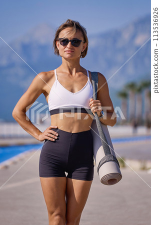 Smiling young sportswoman holding fitness mat, walking outdoors at the seaside Smiling young sportswoman holding fitness mat, walking outdoors at the seaside 113536926