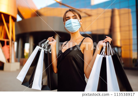 Beautiful woman in protective sterile medical mask with shopping bags near shopping center.Covid- 2019. Beautiful woman in protective sterile medical mask with shopping bags near shopping center.Covid- 2019. 113538917