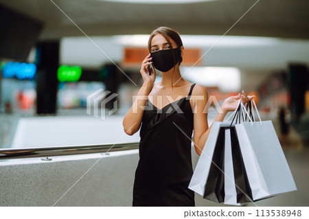 Young woman in protective black medical mask with shopping bags talking on a mobile phone in the mall. 113538948