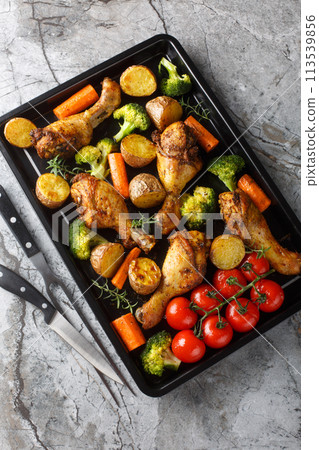 Baked chicken drumstick with broccoli, potatoes, tomatoes, and carrots close-up on a sheet pan. Vertical top view 113539856