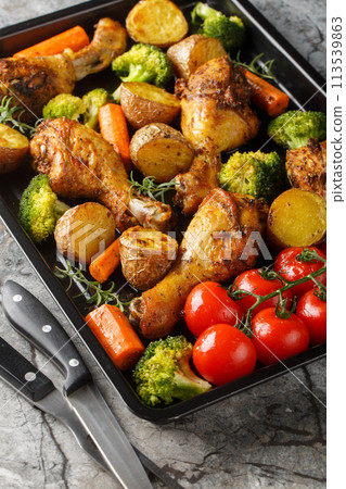 Baked chicken drumstick with broccoli, potatoes, tomatoes, and carrots close-up on a sheet pan. Vertical 113539863