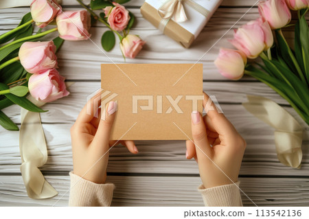 Hands holding blank paper card mockup on wooden desk with flowers and gift box. 113542136