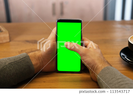 Young man sitting at cafe holding smartphone green mock-up screen in hand. Male person using chroma key mobile phone. Vertical mode. Touching, swiping display, tapping, surfing internet social media Young man sitting at cafe holding smartphone green mock-up screen in hand. Male person using chroma key mobile phone. Vertical mode. Touching, swiping display, tapping, surfing internet social media 113543717
