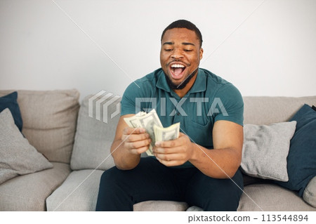 Black man sitting on a coach and holding a cash. Man wearing blue t-shirt. Man posing for a photo and looking very happy. Black man sitting on a coach and holding a cash. Man wearing blue t-shirt. Man posing for a photo and looking very happy. 113544894