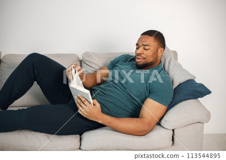 Black man lay on a coach with a book. Man wearing blue t-shirt. Man reading a book at home on a coach. 113544895