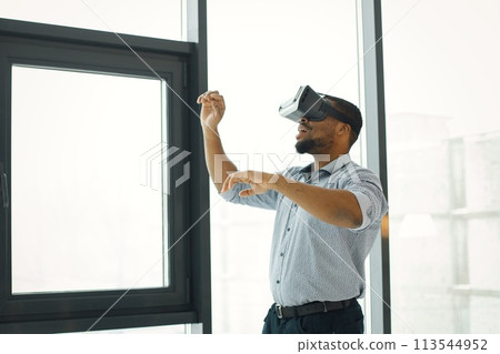 Black business man standing in office near big windows. Bearded man wearing virtual reality glasses. Man wearing blue shirt. Black business man standing in office near big windows. Bearded man wearing virtual reality glasses. Man wearing blue shirt. 113544952