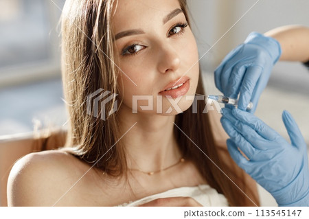 Closeup of doctor's hands in gloves with syringe near female face. Portrait of beautiful caucasian woman with white towel on her bodu. Girl receiving beauty injection. 113545147